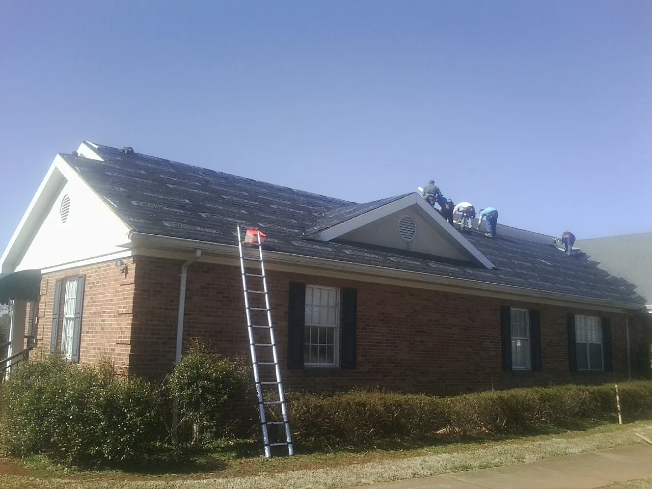Crew tearing shingles off a brick ranch roof with a ladder set — Metal Roof Repair work by our crew in Thompson